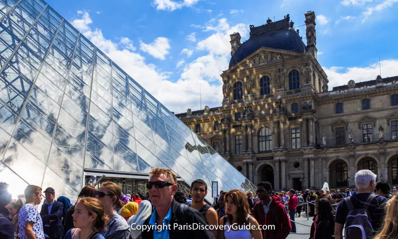 Ticket line at the Louvre winding around the courtyard; nearby signs warn of a 2+ hour wait Ticket line at the Louvre winding around the courtyard; nearby signs warn of a 2+ hour wait