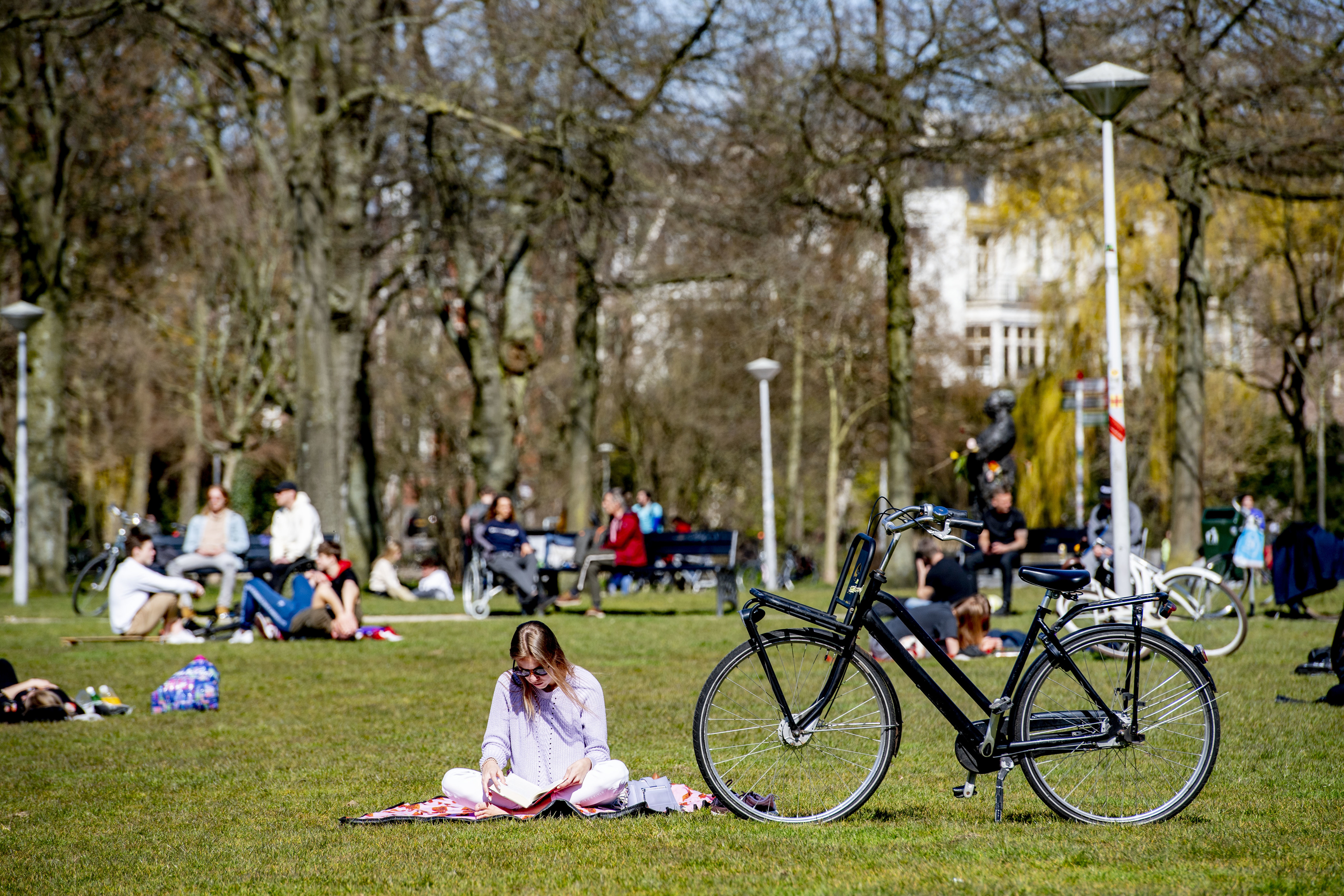 A woman sits cross legged and reads a book in the sunshine during a spring day at the Vondelpark in Amsterdam. She is sat next to her black bicycle with trees and other park users in the background.